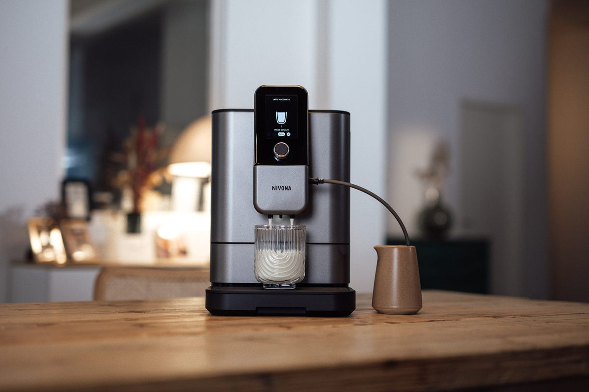 NIVONA NIVO 8'103 auf einem Holztisch im Abendlicht, ein Latte Macchiato läuft ins Glas, daneben ein Milchkännchen